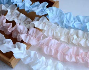 May include: Close-up of ruffled fabric trim in shades of white, light pink, and light blue. The trim is arranged in wavy lines, with the fabric gathered to create a ruffled effect. The trim is likely used for sewing or crafting projects.
