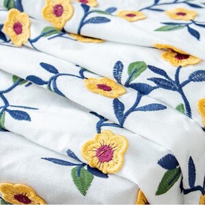 May include: White fabric with a floral embroidery pattern. The flowers are yellow with a burgundy center and are arranged on blue and green stems.