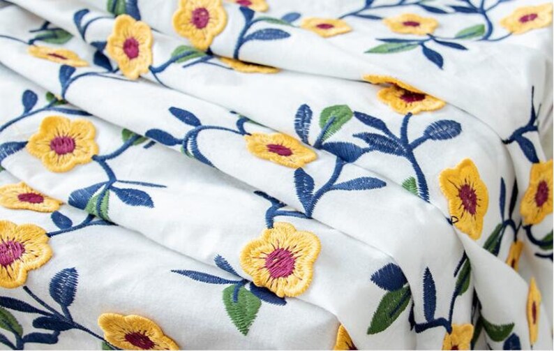 May include: White fabric with a floral embroidery pattern. The flowers are yellow with a burgundy center and are arranged on blue and green stems.