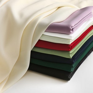 May include: A stack of fabric swatches in various colors, including cream, purple, white, red, green, navy, and dark green. The fabric is smooth and appears to be a lightweight material.