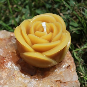 Puede incluir: Una vela de cera de abeja amarilla en forma de rosa se encuentra sobre un grupo de cristales de cuarzo transparentes. La vela tiene forma de rosa con una mecha en el centro.