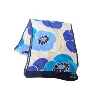 May include: A rectangular scarf featuring a white background with large blue and light blue floral patterns. The design includes thin green stems and a dark blue border at the bottom edge. The word OSCAR is printed in white near the bottom corner.
