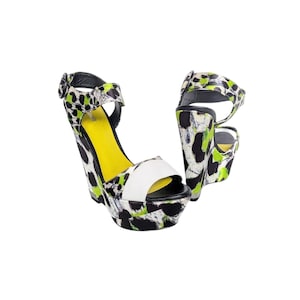 May include: A pair of wedge sandals featuring a black, white, and neon green abstract animal print pattern. The shoes have a white strap across the open toe, a black ankle strap with a buckle, and a bright yellow insole. The platform wedge heel is covered in the same patterned material.