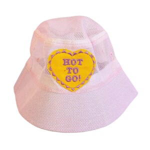 May include: A light pink bucket hat with a mesh crown and brim. A yellow heart-shaped patch with the words "HOT TO GO!" in purple lettering is sewn on the front. The hat is designed for sun protection and casual wear.