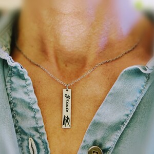 May include: A silver chain necklace with a silver rectangular pendant that says "Gracie" with a silhouette of a person holding a flag.