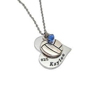 May include: Silver heart-shaped necklace with a volleyball charm and a blue crystal. The heart pendant has the text "#25 Kaylee" engraved on it.