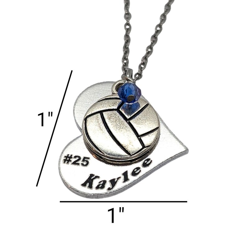 May include: Silver heart-shaped pendant necklace featuring a volleyball charm and a blue crystal. The pendant is engraved with the text "#25 Kaylee" and measures 2.5 cm in diameter.