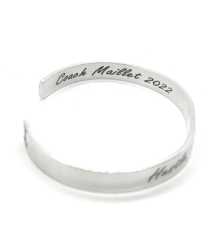 May include: Silver metal bracelet inscribed with "Coach Maillet 2022" on the top and "Hustle" on the bottom.