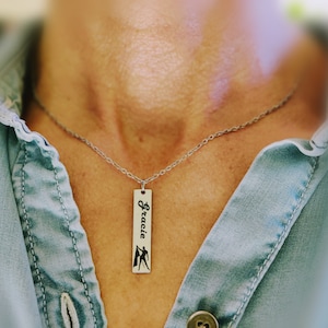 May include: A silver chain necklace with a silver rectangular pendant that says "Gracie" with a silhouette of a person holding a flag.
