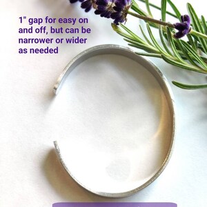 May include: A silver metal bangle bracelet with a 2.5 cm gap for easy on and off. The bracelet is on a white background with lavender flowers and rosemary.