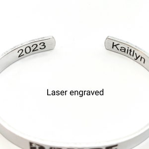 May include: Silver bangle bracelet with laser engraved text "Kaitlyn" and "2023".