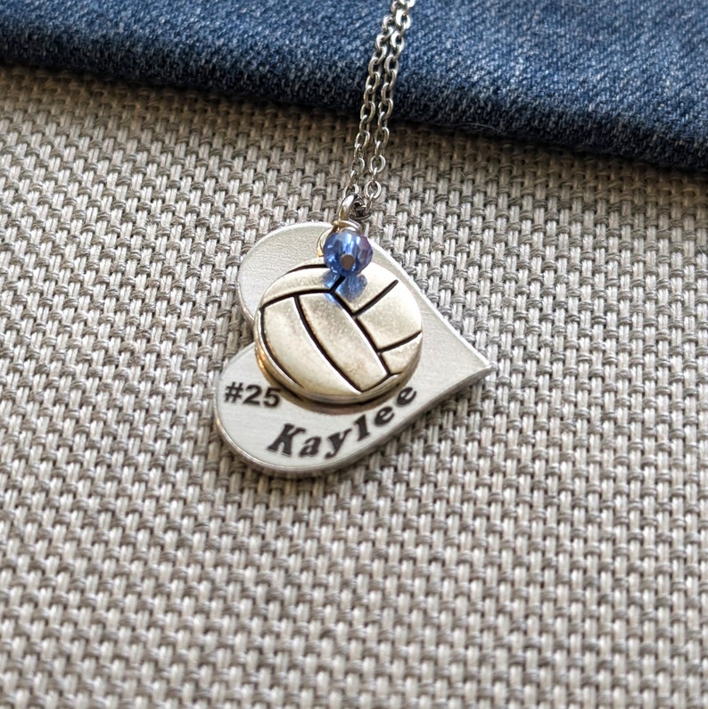 May include: Silver heart-shaped necklace with a volleyball charm and a blue crystal. The necklace is engraved with "#25 Kaylee".