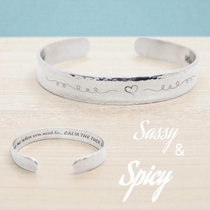 May include: Two silver-colored cuff bracelets. The top bracelet has a hammered texture with a heart and line design. The bottom bracelet has the text "...when you need to... CALM THE FUCK..." The words "Sassy & Spicy" are in the background.