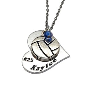 May include: Silver heart-shaped necklace with a volleyball charm and a blue crystal. The heart has the text "#25 Kaylee" engraved on it.