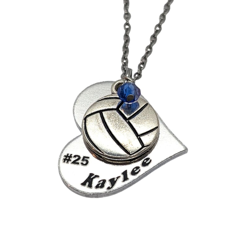 May include: Silver heart-shaped necklace with a volleyball charm and a blue crystal. The heart has the text "#25 Kaylee" engraved on it.