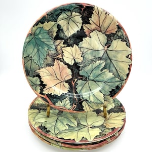 May include: A stack of three round ceramic plates featuring a detailed botanical pattern of overlapping grape leaves in shades of sage green, cream, and dark brown. The edges of the plates are finished with a thin, warm reddish-brown rim.