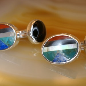 May include: A pair of oval-shaped cufflinks with a silver setting. Each cufflink features a multi-colored inlay of red, black, white, turquoise, blue, and lapis lazuli stones. The other side of each cufflink is black.