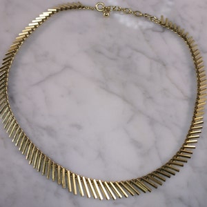 May include: A gold necklace with a geometric design made of small, rectangular pieces. The necklace is on a white marble surface.