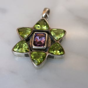 May include: A silver pendant in the shape of a six-pointed star with a purple gemstone in the center and five green gemstones around it.