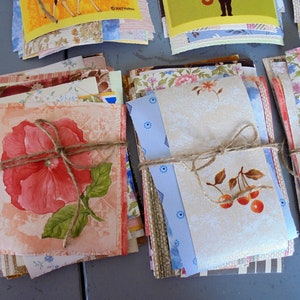 Könnte beinhalten: Ein Stapel Vintage-Papierreste mit floralen und geometrischen Mustern, zusammengebunden mit Schnur. Der oberste Rest zeigt eine rosa Blume mit grünen Blättern. Die Reste sind perfekt zum Basteln, Scrapbooking oder Decoupage.