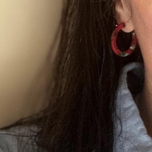 May include: Red and green patterned hoop earrings. The earrings feature a vibrant red hue with a speckled green and white design. These hoop earrings are a classic style, suitable for everyday wear or special occasions.