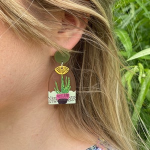 May include: A pair of green and yellow resin earrings featuring a potted plant with a pink pot and a yellow flower on top. The earrings are shaped like an arch.