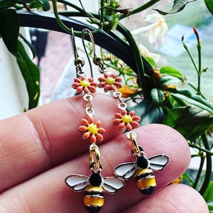 May include: Gold-coloured earrings with a bee and flower design. The earrings have a yellow, black, and white bee charm, and a chain of orange and yellow flower charms. The earrings are suspended from a gold-coloured hook.