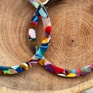 May include: A pair of large, colourful hoop earrings made of resin. The earrings are a mix of blue, red, yellow, green, and white.