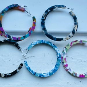 May include: Five pairs of hoop earrings in a marbled design. The earrings feature a variety of colours, including blue, black, pink, and green. They are made from a lightweight material and have a post closure.
