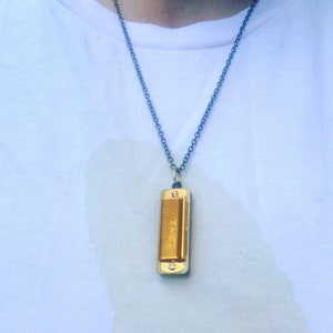Könnte beinhalten: Eine goldene Mundharmonika-Kette mit einer schwarzen Kette. Die Mundharmonika hat den Schriftzug "Hohner" aufgedruckt.