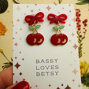 May include: A pair of cherry earrings with red bows and green leaves. The earrings are on a white card with the text "BASSY LOVES BETSY". The background features botanical illustrations and a pattern of diamond shapes.