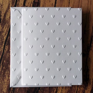 May include: White stationery set featuring a textured heart pattern. The set includes a rectangular card and an envelope, both with a repeating embossed heart design. The paper has a slightly raised texture, creating a subtle, tactile effect. The background is a wooden surface.