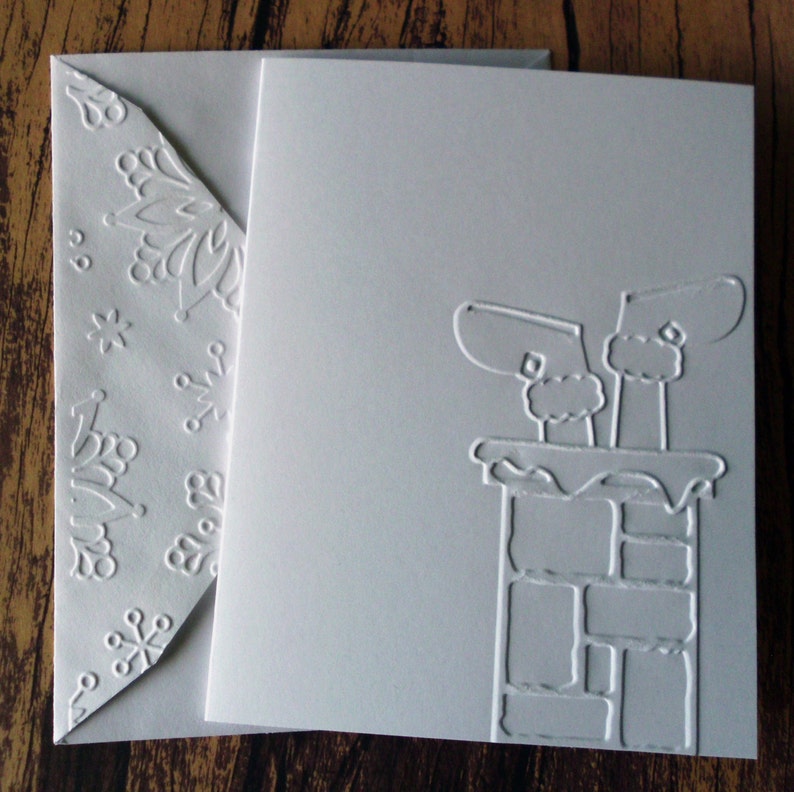 May include: A white Christmas card with an embossed design of Santa's boots in a chimney. The card is accompanied by a matching envelope with a snowflake pattern. The card and envelope are made of thick, textured paper.