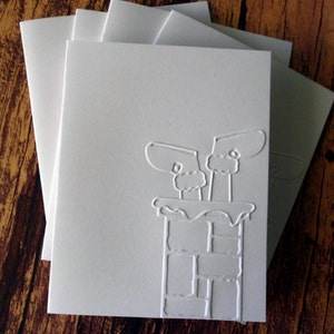 May include: A stack of white greeting cards with embossed chimney and mittens design. The cards are blank inside and have matching envelopes. The design is a winter holiday theme.
