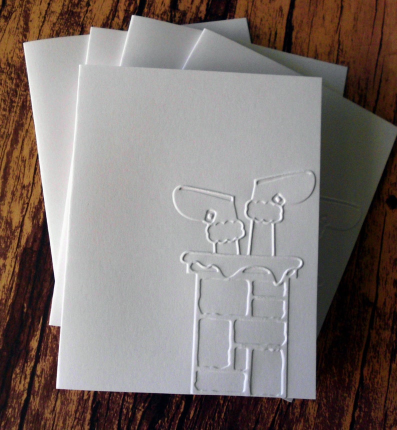 May include: A stack of white greeting cards with embossed chimney and mittens design. The cards are blank inside and have matching envelopes. The design is a winter holiday theme.