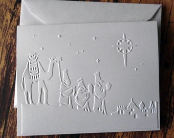 May include: A white embossed Christmas card with a scene of the three wise men following a star on a camel. The card has a white background and the image is embossed in white.
