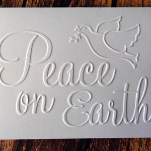 Pu&ograve; includere: Biglietto bianco in rilievo con le parole "Peace on Earth" in elegante scrittura. &Egrave; presente anche un'immagine in rilievo di una colomba con un ramoscello d'ulivo, creando un effetto strutturato tridimensionale. Il biglietto &egrave; su uno sfondo di legno.