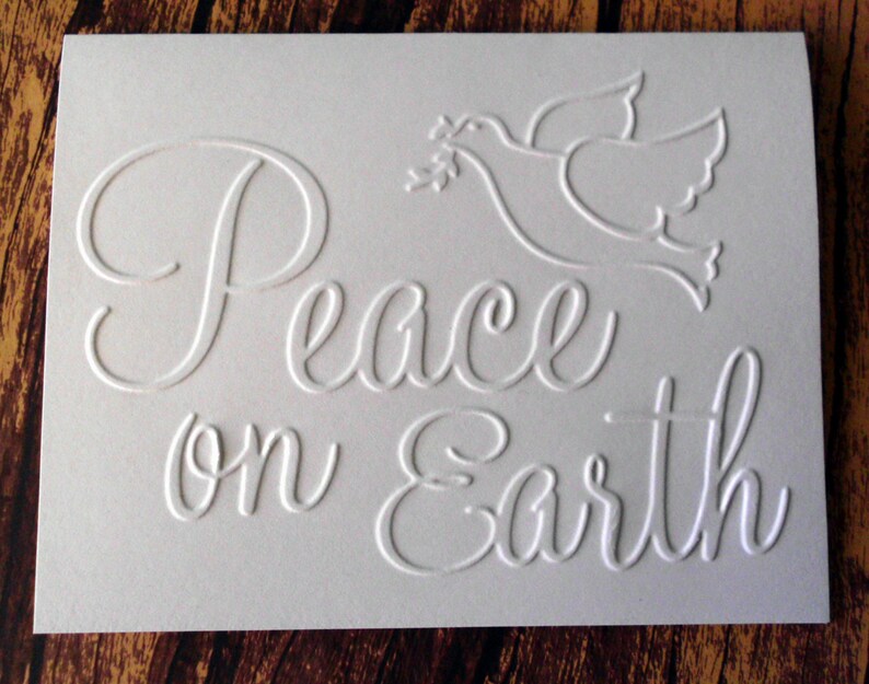 Pu&ograve; includere: Biglietto bianco in rilievo con le parole "Peace on Earth" in elegante scrittura. &Egrave; presente anche un'immagine in rilievo di una colomba con un ramoscello d'ulivo, creando un effetto strutturato tridimensionale. Il biglietto &egrave; su uno sfondo di legno.