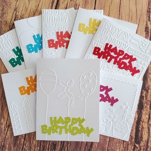 May include: A set of eight white greeting cards with embossed designs and colorful paper cutouts. The cards feature the words "Happy Birthday" in various fonts and colors. The cards are perfect for celebrating a birthday.