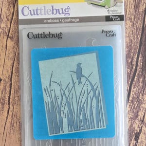 May include: A Cuttlebug Bird Call embossing folder in its original packaging. The clear plastic folder features a cutout design of a bird perched on tall grass. The packaging includes the Cuttlebug logo, Provo Craft branding, and a small illustration of the machine.