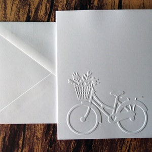 May include: White card with a white embossed image of a bicycle with a basket of flowers. The card has a white envelope.
