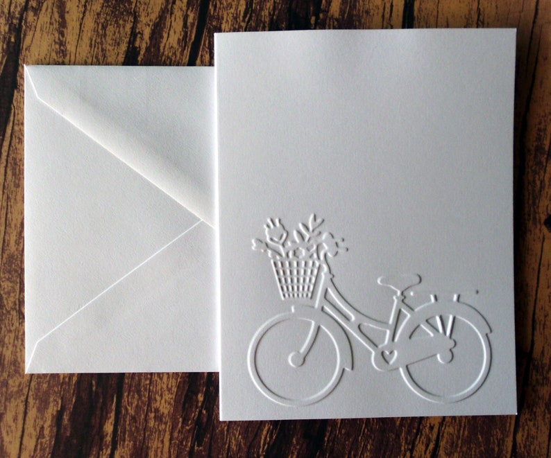 May include: White card with a white embossed image of a bicycle with a basket of flowers. The card has a white envelope.