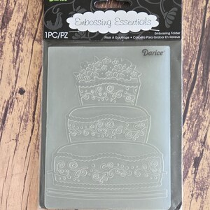 May include: A Darice Embossing Essentials embossing folder in its original black packaging. The clear plastic folder features a three-tiered cake design with floral and swirl patterns. The packaging includes the text Embossing Essentials and 1PC/PZ.