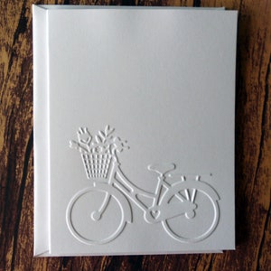 May include: A white card with an embossed image of a bicycle with a basket of flowers. The bicycle has a heart-shaped design on the frame.