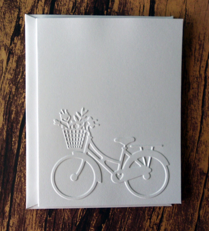 May include: A white card with an embossed image of a bicycle with a basket of flowers. The bicycle has a heart-shaped design on the frame.