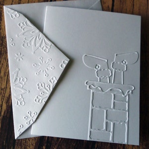 May include: A white Christmas card with an embossed design of Santa's boots in a chimney. The envelope features embossed snowflakes. The card and envelope are made of thick, textured paper.