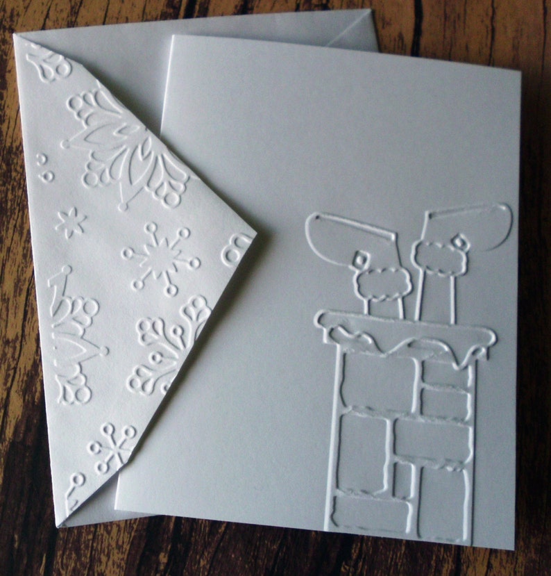 May include: A white Christmas card with an embossed design of Santa's boots in a chimney. The envelope features embossed snowflakes. The card and envelope are made of thick, textured paper.
