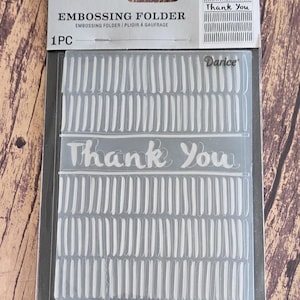 May include: A clear plastic Darice embossing folder in its original retail packaging. The folder features a repeating pattern of vertical dashed lines and the text Thank You centered horizontally. The packaging includes the brand name Darice.