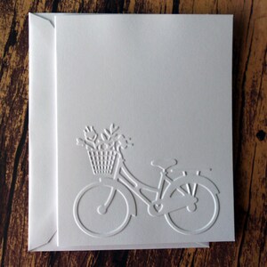 May include: White card with an embossed image of a bicycle with a basket of flowers. The bicycle is facing right and has a heart-shaped design on the frame.