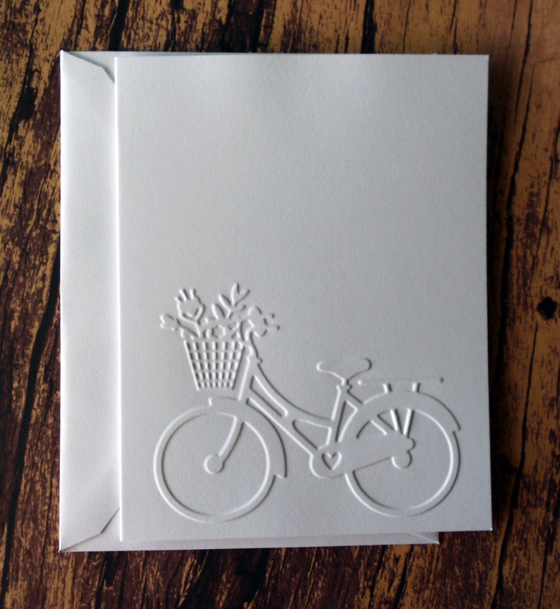 May include: White card with an embossed image of a bicycle with a basket of flowers. The bicycle is facing right and has a heart-shaped design on the frame.